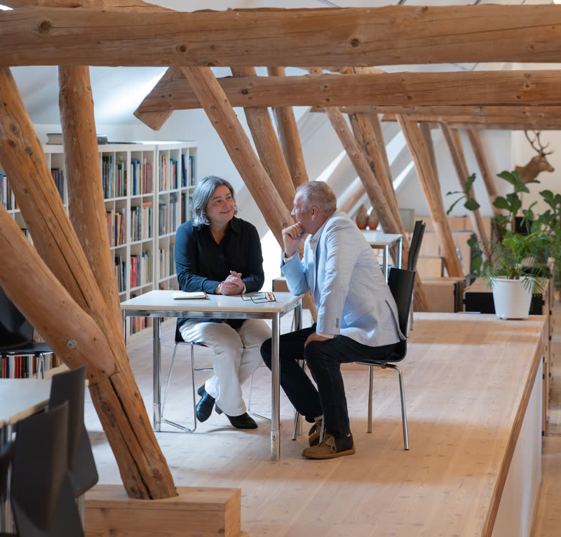 To personer sidder i samtale ved et bord i et loftsrum med synlige tømmerbjælker.