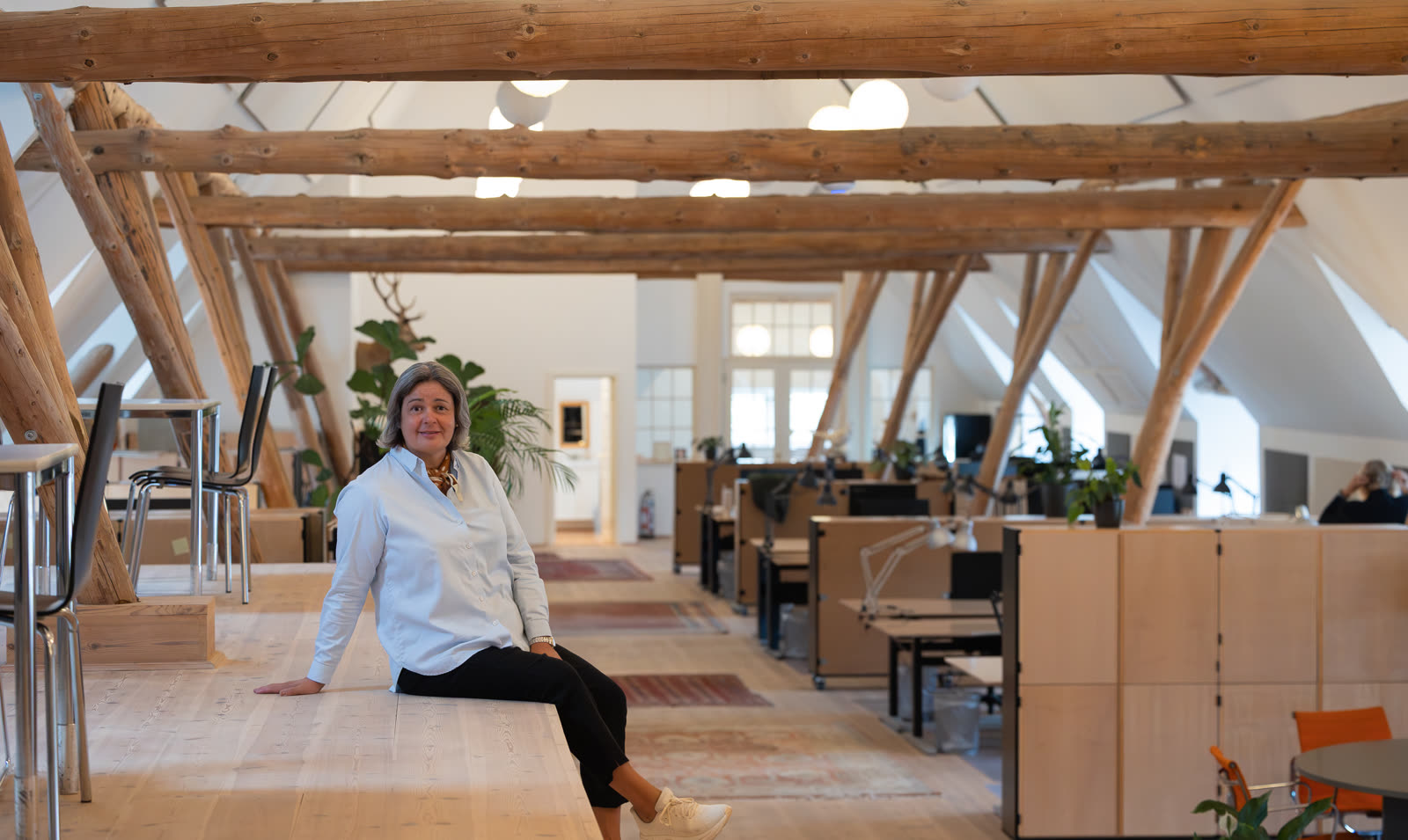 Lone Rosenørn Jakobsen sidder på et langbord i et lyst kontorlokale med synlige tømmerbjælker.